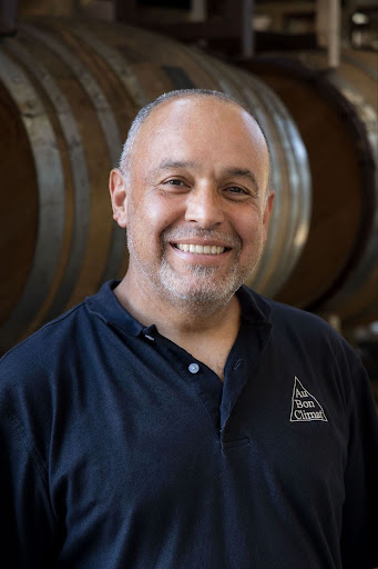 A photo of Au Bon Climat's cellarmaster, Enrique smiling as he stands in front of wooden wine barrels.