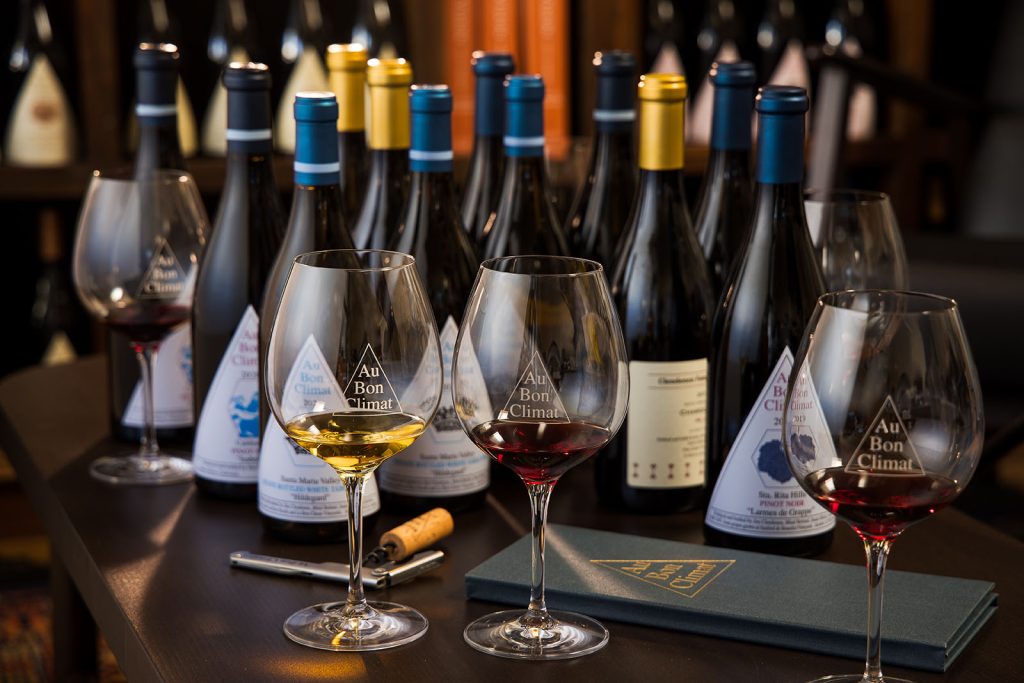 Red and white wine in Au Bon Climat wine glasses with many bottles behind.