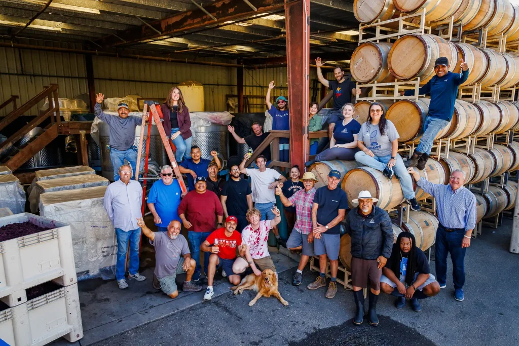 Group photo of the Au Bon Climat harvest team gathered in the winery among barrels and bins.