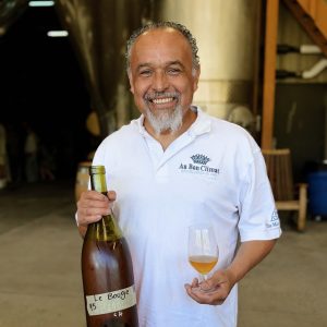Enrique stands before the production facility and wine tanks smiling with an unlabeled bottle of wine and glass.