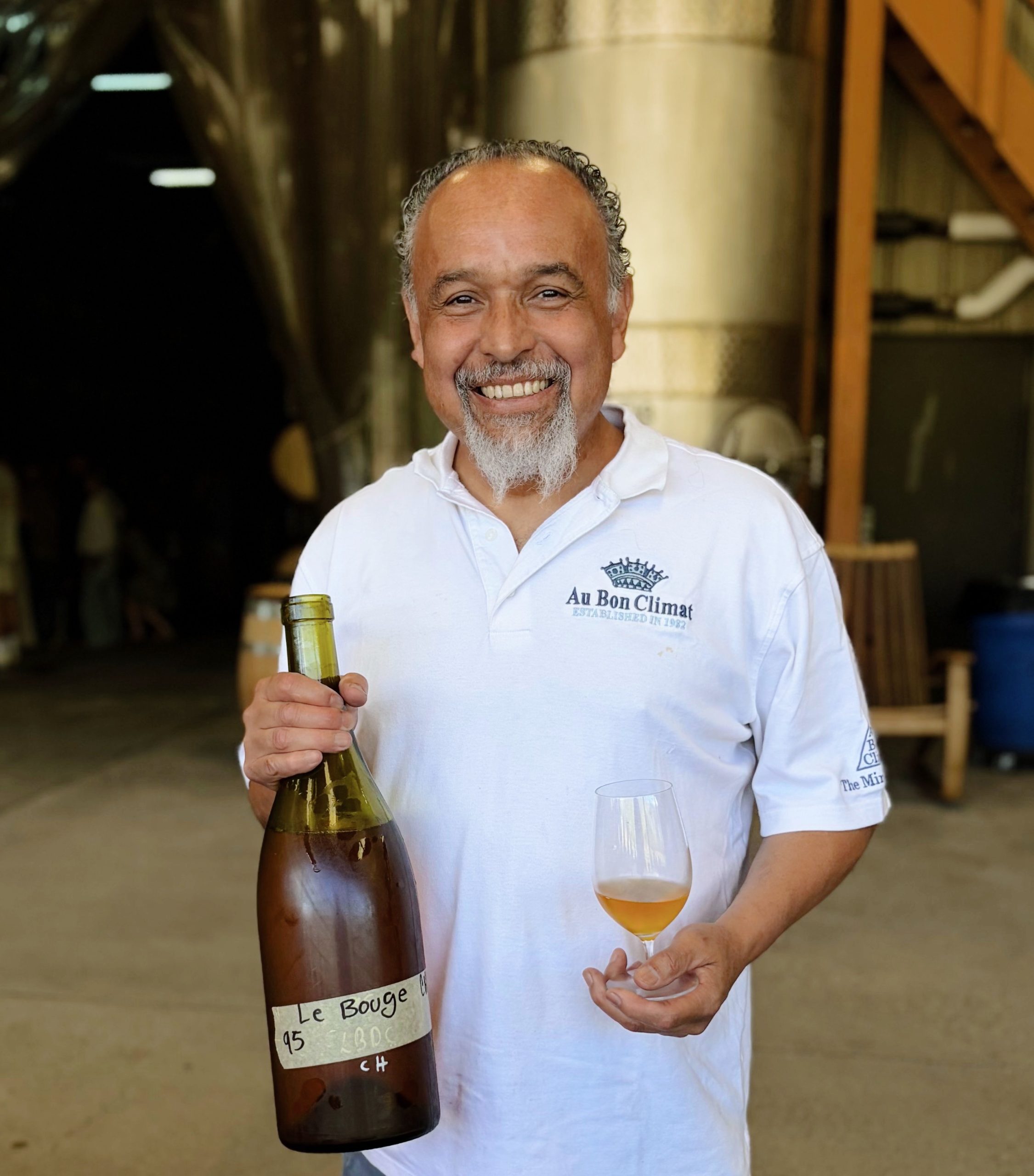 Enrique stands before the production facility and wine tanks smiling with an unlabeled bottle of wine and glass.